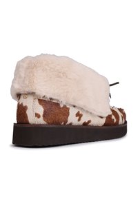 Chausson en imprimé vache marron et crème avec un dessus en fausse fourrure, semelle plate marron et un détail en dentelle à l'avant. Texture douce et forme arrondie.