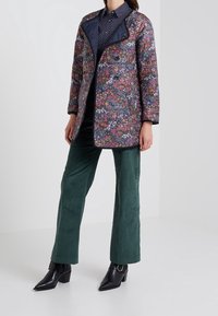 Kukkaiskuvioitu, vedenpitävä takki, jossa on tummansininen kaulus. Yhdistetty vihreisiin puuvilla-kanaalihaalareihin ja mustiin kärjellisiin nilkkabootseihin.