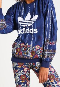 Felpa blu navy con logo Adidas bianco, motivi floreali intricati sull'orlo e sulle maniche, abbinata a leggings con motivo coordinato.
