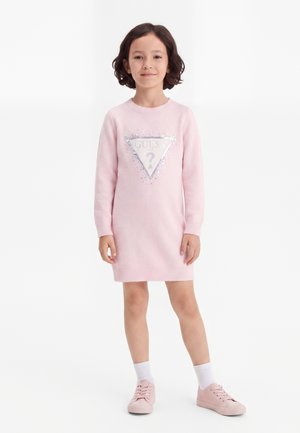 Barn står iført en lys rosa langærmet kjole med en sølv trekantet logo og lyserøde sko med hvide sokker på hvid baggrund.