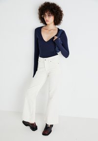 Tengerész navy hosszúujjú bodysuit, fehér trapézfarmer, és vastag, két színű cipők. A modell egy texturált kiegészítőt tart, göndör haja és szemüvege van.