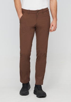 Homme portant un pantalon slim marron et des baskets noires, debout avec une main dans la poche sur un fond clair uni.
