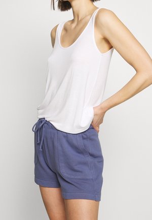 Frau trägt ein weißes ärmelloses Tanktop und blaue Shorts mit Kordelzug, steht mit einer Hand in der Hüfte vor einem einfarbigen Hintergrund.