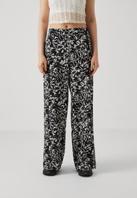 Osoba oblečená v kalhotách s abstraktním černobílým potiskem a širokými nohavicemi, bílém krajkovém crop topu a černých teniskách se žlutými detaily.