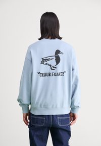 Sudadera azul claro de cuello redondo con un gráfico de un pato negro y la palabra "TROUBLEMAKER" en la parte posterior. Material suave de mezcla de algodón.
