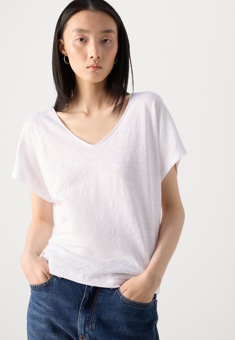 Biała koszulka z krótkim rękawem w serek, wykonana z lekkiego, lekko teksturowanego materiału. Połączona z niebieskimi dżinsami, ręce w kieszeniach.