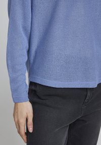 Pull en tricot bleu clair, texturé avec un tissage lâche. Manches longues, ourlet légèrement court, porté sur un jean noir.