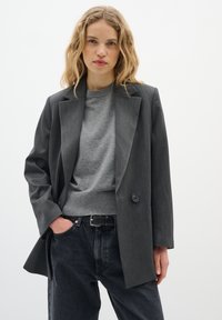 Grå dobbeltradet blazer med skræddersyet pasform, der har strukturerede skuldre, en enkelt sort knap, båret over en grå sweater og mørke jeans.