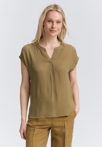 Blouse vert olive avec un col mandarin, des manches courtes et une coupe ample. Tissu lisse avec un léger éclat, sans motifs ni accents.