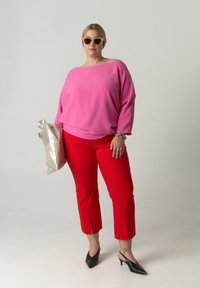 Blouse rose surdimensionnée, pantalon rouge cropped, escarpins noirs à bout pointu. Tenant un sac argenté brillant. Tissu lisse, coupe décontractée.