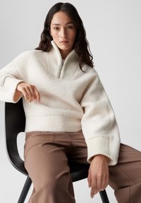 Cremefarbener Fleece-Pullover mit hohem, geripptem Kragen und Reißverschluss, ausgestattet mit weiten Ärmeln. Kombiniert mit braunen, maßgeschneiderten Hosen.