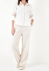 Camicia bianca in lino con ricami decorativi sulle maniche, abbinata a pantaloni beige a gamba larga e scarpe chiare.