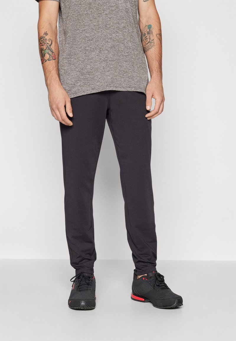 Pantalon de survêtement noir avec une coupe fuselée, fabriqué à partir d'un tissu doux. Porté avec un t-shirt gris et des chaussures de sport noires avec des accents rouges.