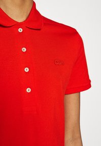 Gros plan sur une personne portant un polo rouge vif avec des boutons blancs et un logo de crocodile rouge brodé sur la poitrine.