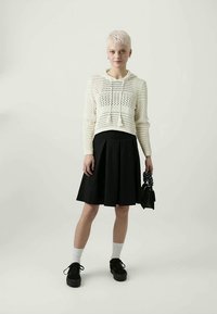 Felpa corta lavorata a maglia bianca con coulisse, gonna plissettata nera, calze bianche, scarpe con plateau nere e una piccola borsa nera.