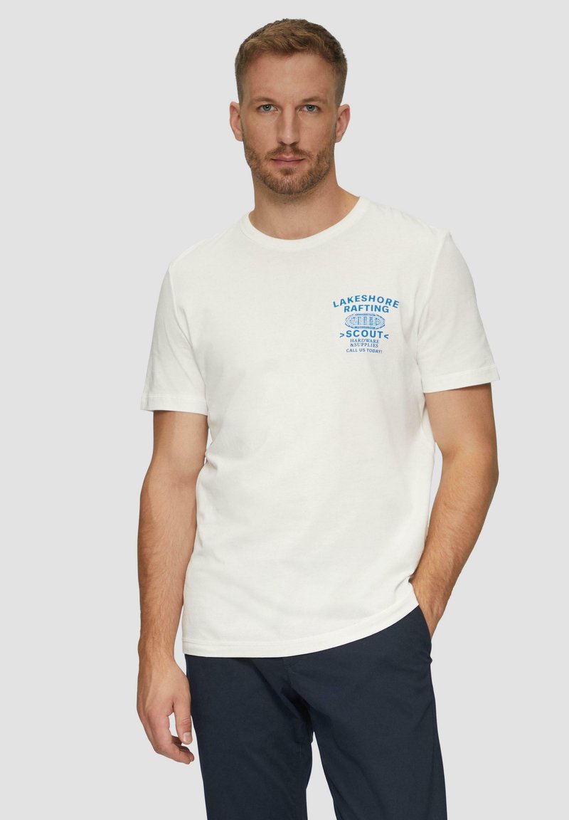 Witte katoenen t-shirt met blauwe grafische tekst op de linkerborst. Korte mouwen en ronde hals. Draagt het met marineblauwe broek.