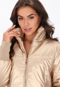 Giacca puffer beige con colletto alto, chiusura con zip e polsini a costine. Il tessuto ha una consistenza liscia e lucida e presenta un accento dorato.