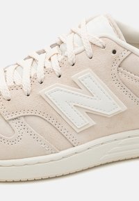 Baskets en daim beige avec un logo "N" blanc à grosse couture sur le côté, lacets crème et col rembourré sur une semelle en caoutchouc blanche.