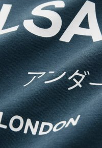 Jūrų laivyno audinys su tekstūruotu audimu, kuris pasižymi ryškiu baltu tekstu. Tekste įtrauktas žodis "LONDON" ir japonų simboliai, kurie išryškinti.
