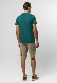 Īsām piedurknēm teal krāsas t-krekls savienots ar khaki krāsas šortiem. Šortiem ir sānu kabatas. Modelis valkā brūnas čības un ir vērsts prom.