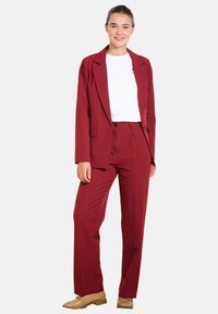 Burgundski blazer in ujemajočih se hlačah iz gladkega materiala, ki vključuje žepke z flapo na blazernem modelu in sproščen kroj, nosi pa ga z belo srajco.