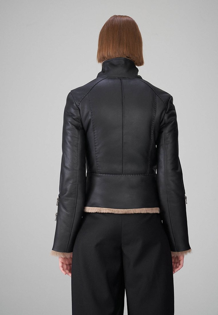 Schwarze Lederjacke mit hohem Kragen und tailliertem Design, versehen mit weichem beigem Pelzbesatz am Saum und an den Ärmelbündchen. Reißverschlüsse an den Ärmeln.