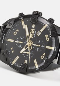 Montre en acier inoxydable noir avec un boîtier rond, des accents dorés, des sous-cadrans chronographe et des marqueurs d'heure audacieux. Résistante à l'eau jusqu'à 5 bars.