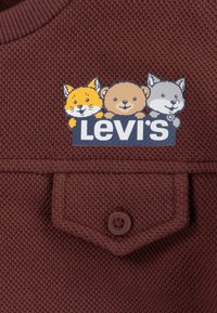 Brun texturerad tyg med en framknäppt ficka och en färgglad Levi's-logotyp som har tecknade ansikten av en räv, björn och varg ovanför.