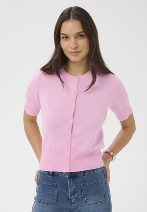 Hosszú sötét hajú nő, rózsaszín, rövid ujjú gombos kardigánt és kék farmert visel, kezét a háta mögött tartva áll.