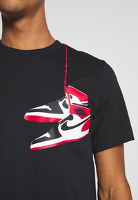 Camiseta negra con un diseño gráfico de zapatillas rojas y negras con acentos blancos, destacadas de forma prominente en la tela.