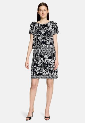 Frau steht und trägt ein schwarz-weißes, floral gemustertes Kurzarmkleid mit geometrischem Besatz und schwarze offene High Heels vor weißem Hintergrund.