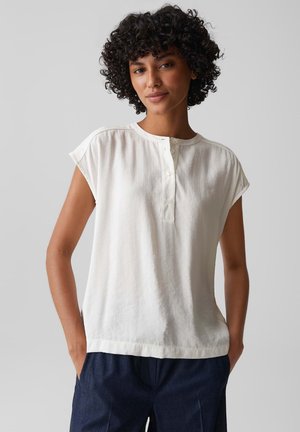 Femme aux cheveux bouclés portant un chemisier blanc à manches courtes boutonné et un pantalon bleu foncé, debout les mains dans les poches devant un fond uni.
