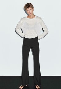 Lichtgrijs, semi-doorzichtig longsleeve shirt gecombineerd met zwarte hoge taille flared broeken; blootvoets gedragen tegen een eenvoudige achtergrond.