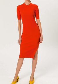 Femme portant une robe ajustée, côtelée, orange, longueur genoux, avec des manches courtes et une fente latérale, associée à des chaussures à talons hauts jaunes.