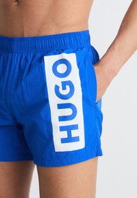 Shorts de bain bleus en matériau léger avec un panneau vertical blanc portant le texte bleu "HUGO", taille élastique et poche latérale.
