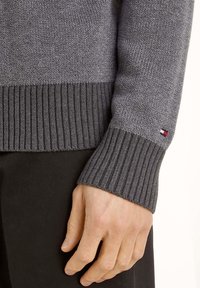 Grau gestrickter Pullover mit gerippten Bündchen an Ärmeln und Saum. Auf dem Ärmel befindet sich ein kleines Logotag. Das Material weist eine strukturierte Oberfläche auf.