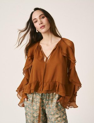 Mujer de cabello largo y oscuro lleva una blusa color óxido con volantes y pantalones verdes de pierna ancha con estampado, de pie con las manos en los bolsillos.