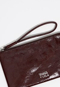 Burgundska teksturirana usnjena denarnica z ročajem za zapestje, z srebrno logotipno ploščico. Zadrga na vrhu. Gladek, sijoč zaključek.