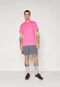 Rosa Sport-T-Shirt, graue Shorts, schwarze Laufschuhe und graue Socken. Zeigt Logos auf den Kleidungsstücken und sichtbare Tätowierungen an den Armen.