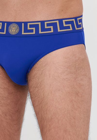 Blaue Schwimmslips mit einem goldenen Bund im griechischen Schlüsselmuster und einem zentralen Medusa-Emblem. Glatte Textur und figurschmeichelndes Design.