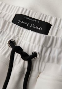 Białe szorty z czarnym sznurkiem, elastycznym paskiem w talii oraz metalowymi oplotami. Metka mówi "Ombre Eximio" z napisem "Premium Quality".