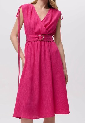 Mujer con un vestido midi texturizado de color rosa brillante con escote en V, sin mangas, tirantes anudados en los hombros y un cinturón a juego con hebilla metálica.
