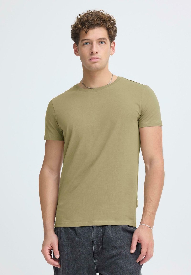 Junger Mann mit lockigem Haar, der ein olivgrünes eng anliegendes T-Shirt und dunkle Jeans trägt, vor einem schlichten hellen Hintergrund stehend.