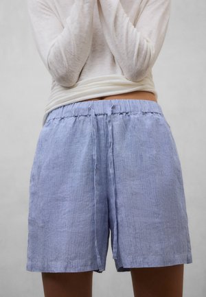 Person trägt hellblaue und weiß gestreifte Shorts mit Kordelzug und ein langärmeliges weißes Oberteil, die Arme vor der Brust verschränkt.