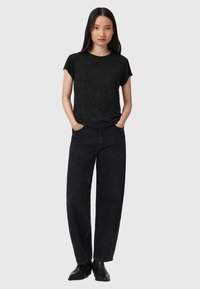 Mujer con cabello lacio negro, vistiendo una camiseta negra con manchas, jeans negros holgados y botas de tobillo negras, de pie con las manos en los bolsillos.
