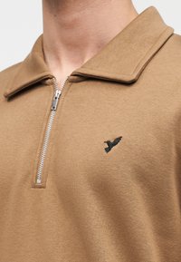 Καφέ πόλο με φερμουάρ, με φαρδιά γιακά και μικρό μαύρο έμβλημα πουλιού στο αριστερό στήθος, κ ατασκευασμένο από απαλό, υφή υφάσματος.