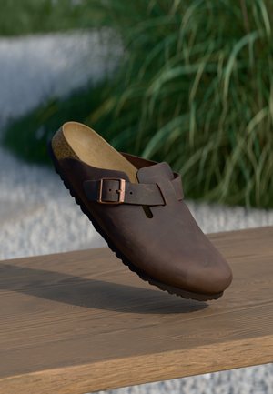 Brauner Lederclog mit Aussparung, verstellbarem Riemen und Kupfer-Schnalle sowie strukturiertem Kork-Fußbett, erhöht über einer Holzoberfläche.