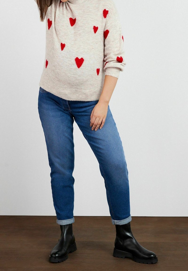Personne portant un jean bleu retroussé, des bottes noires et un pull beige avec des motifs de cœurs rouges, debout sur un sol en bois.