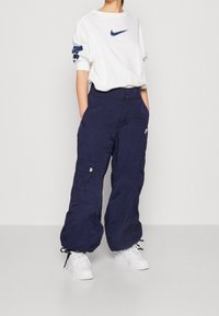 Marine cargobroek met zijzakken en verstelbare manchetten, gecombineerd met een wit sweatshirt met een marine Nike-logo. Sneakers maken de look af.