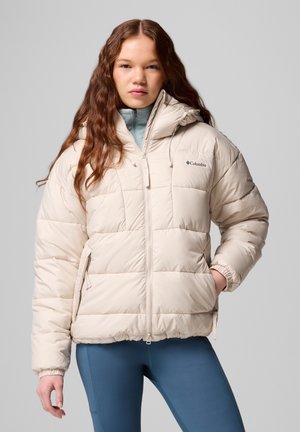 Joven mujer con largo cabello rojo, vistiendo una chaqueta acolchada beige claro y leggings azules, con las manos en los bolsillos de la chaqueta, mirando hacia adelante.